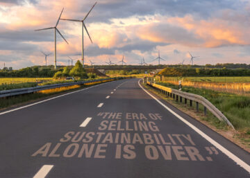 a paved two-way road with open fields on both sides and wind turbines at a distance