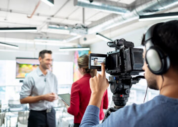 Two people talking in a bright office space. A man recording their conversation checks his camera monitor.