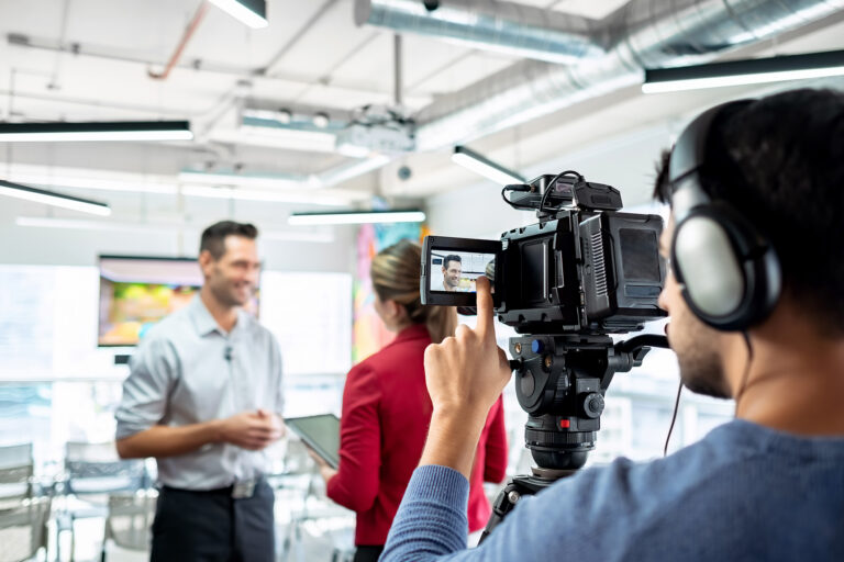 Two people talking in a bright office space. A man recording their conversation checks his camera monitor.